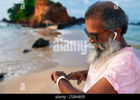 Senior Ethno Mann verwendet Smart Watch und kabellose Ohrstöpsel am tropischen Strand. Bärtige ältere Menschen genießen moderne Technik für Fitness und Wohlbefinden. Gesunder Lebensstil Stockfoto