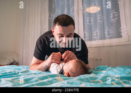 Ein hingebungsvoller Vater lehnt sich auf einer weichen blauen Decke über sein Baby, während er sanft lächelt, während er die kleinen Bewegungen seines Kindes in einem weißen Einteiler beobachtet. Stockfoto