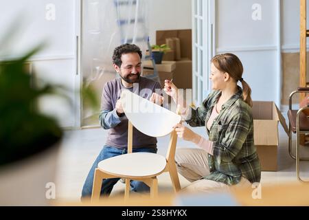 Glückliche junge Frau, die ihrem Mann Nägel gibt, um den neuen Stuhl zu reparieren, während beide im Wohnzimmer auf Hocken stehen Stockfoto