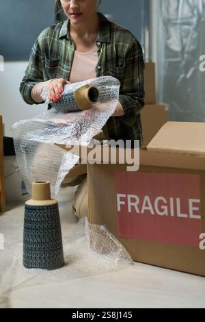 Abgeschnittener Schuss einer jungen Frau, die auf Squats neben einer offenen Box sitzt und die Vase vor dem Packen in Luftpolsterfolie wickelt Stockfoto