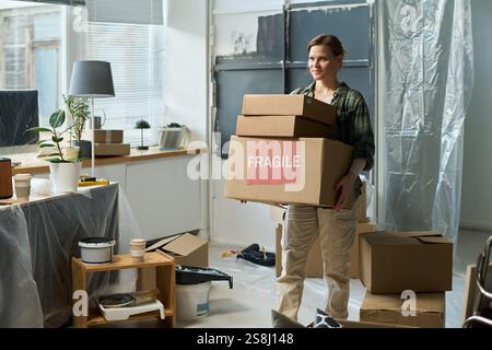 Junge Frau in lässiger Kleidung, die Kartons mit zerbrechlichen Haushaltssachen trägt, während sie durch das Wohnzimmer läuft Stockfoto