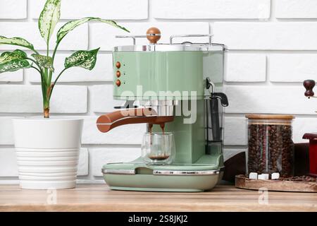 Moderne Kaffeemaschine mit einem Glas heißem Espresso und einem Glas Bohnen auf einem Holztisch in der Nähe der weißen Backsteinwand in der Küche Stockfoto