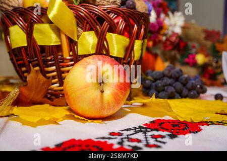 Ein lebhafter Herbstvortrag auf einem Tisch zeigt einen frischen Apfel, Traubensträhne, bunte Blätter und einen Korb mit saisonalen Dekorationen. Stockfoto