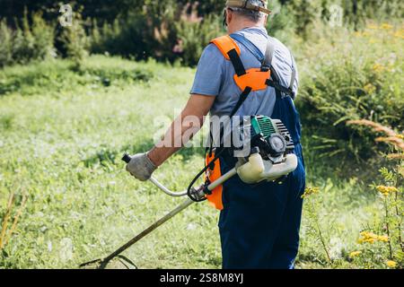 Senior man mäht das Gras mit einem Trimmer an klaren Tagen hinter dem Haus. Stockfoto