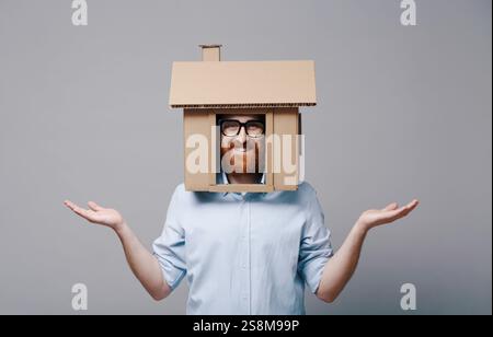 Lächelnder, selbstbewusster Mann mit Kopf in einem Papphaus: Immobilien-, Wohn- und Versicherungslösungen Stockfoto