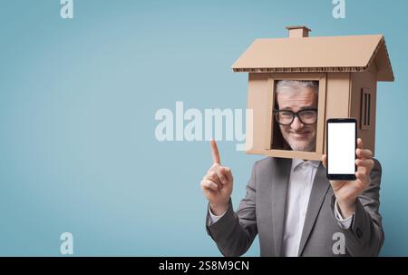 Ein lächelnder Immobilienmakler oder Versicherungsmakler mit Kopf in einem Papphaus zeigt ein leeres Smartphone Stockfoto