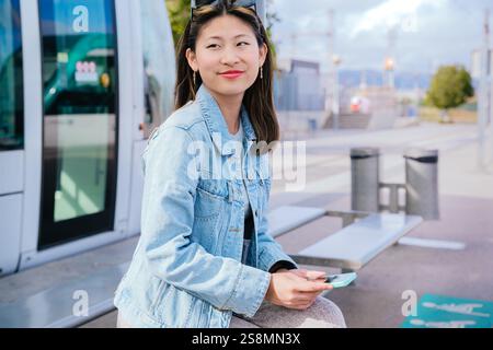 : Junge Frau, die an der Straßenbahnhaltestelle sitzt und ihr Smartphone in der Stadt hält Stockfoto
