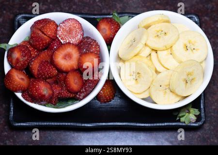 In Scheiben geschnittene Erdbeeren und Bananen auf weißen Schüsseln auf einem schwarzen Teller Stockfoto