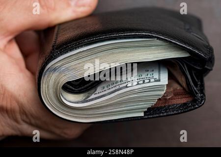 Die Hand eines Mannes hält eine schwarze Ledertasche mit einem Haufen 100-Dollar-Scheine, Banknoten, Bargeld, Nahaufnahme. Konzept für verlorenes oder gefundenes Wallet mit CA Stockfoto