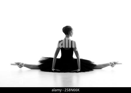Eine professionelle Ballerina in einem schwarzen Tutu führt eine ausgeglichene und anmutige Bodendehnung durch, die als künstlerische schwarz-weiße Silhouette festgehalten wird. Stockfoto
