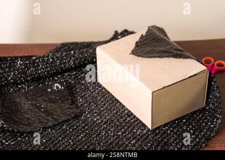 Eine Pappschachtel auf einem Holztisch, teilweise mit schwarzer Luftpolsterfolie bedeckt. Scheren sind in der Nähe sichtbar, was auf die Vorbereitung der Verpackung hindeutet Stockfoto