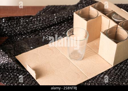 Ein klares Glas auf einer Pappoberfläche, umgeben von einer Schachtel mit weiteren Gläsern. Der Hintergrund ist mit schwarzer Luftpolsterfolie versehen, was auf Packag hindeutet Stockfoto