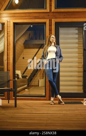 Eine schöne Brünette steht abends am Eingang einer großen Holzhütte Stockfoto
