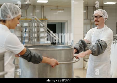 Männer in weißen Uniformen und Haarnetzen bereiten in einer Industrieküche Lebensmittel vor. Ein Arbeiter rührt einen großen Topf, während der andere beobachtet, umgeben von Edelstahlgeräten Stockfoto