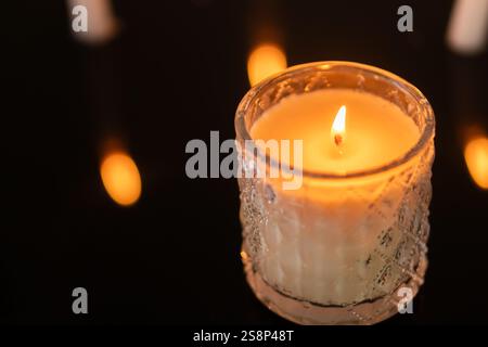 Eine Nahaufnahme einer zündeten Kerze in einem dekorativen Glashalter, mit einem verschwommenen Hintergrund anderer Kerzen. Das warme Leuchten der Flamme erzeugt eine gemütliche Atmosphäre Stockfoto