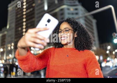 Eine glückliche junge Frau mit lockigem Haar und Brille macht nachts ein Selfie mit ihrem Smartphone in einer Stadt Stockfoto