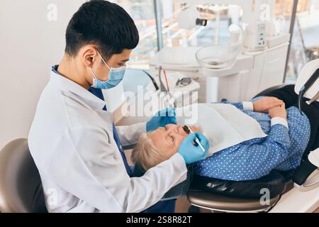 Zahnarzt, der den Mund einer älteren Frau während des zahnärztlichen Eingriffs kontrolliert, Handschuhe mit Hygiene sicherstellt, Patient entspannt auf Stuhl in einer gut beleuchteten Klinik. Stockfoto