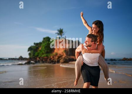 Fröhliche Frau genießt Huckepack-Fahrt auf dem Rücken des Mannes, Paar am Strand bei Sonnenuntergang, verspieltes Date am Meer, Liebe, lustige romantische Momente, Urlaub zusammen Stockfoto