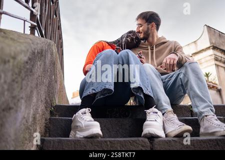 Romantisches, interrassisches Paar, das auf der Treppe der Stadt sitzt und einen zarten Moment zusammen genießt, symbolisiert Liebe, Vielfalt und Verbundenheit in einer urbanen Umgebung. Stockfoto