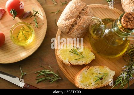 Brotscheiben, gewürzt mit Rosmarinöl auf Holzteller mit Rosmarinzweigen und Tomaten auf einem Holztisch. Draufsicht. Stockfoto