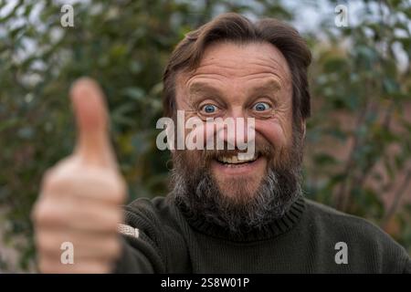 Porträt eines fröhlichen optimistischen Seniorenmannes, der mit dem Schwanz oben in die Kamera schaut. Ältere bärtige kaukasische Männer im Freien im Stadtpark im Ruhestand, Stockfoto