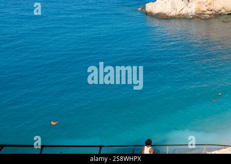 Nicht erkennbare gebräunte Frau, die zum Katsiki Strand in Lefkada - Griechenland absteigt. Das Wasser hat eine tiefe türkisblaue Farbe. Strandgänger im Meer. Stockfoto