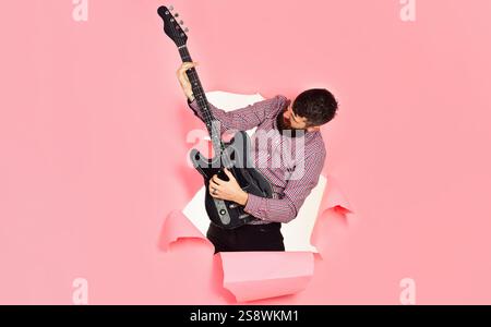 Gitarrenspieler. Bärtiger Mann mit elektrischer Gitarre durch ein Loch in Papier. Rockstar Gitarrist mit E-Gitarre. Hobby, Musikinstrument und Unterhaltung Stockfoto