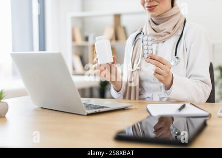 Muslimische Ärztin demonstriert Medikamente während der virtuellen Konsultation mit dem Patienten. Eine Frau aus dem Nahen Osten verwendet Laptop, um Pillen als Behandlung anzubieten, ein Beispiel für Telemedizin. Stockfoto