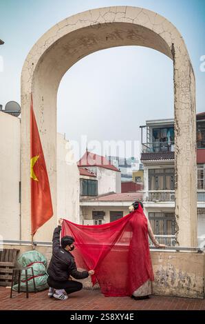 Ein junger Mann hilft seiner Freundin, in ihrem traditionellen ao dai Kostüm auf dem Dach des Dong Xuan Markts in der Altstadt von Hanoi, Vie für Fotos zu posieren Stockfoto