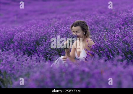 Eine Frau sitzt auf einem Feld aus violetten Blumen und hält eine Lavendelblume Stockfoto