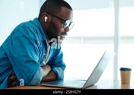 Ein Geschäftsmann nimmt an einem virtuellen Meeting im Büro Teil. Er trägt Ohrhörer und ein lässiges Denim-Hemd, während er sich intensiv auf seinen Laptop konzentriert Stockfoto