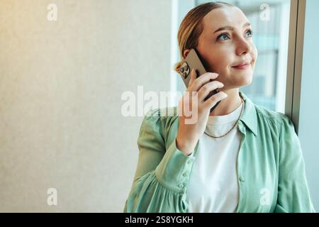 Eine Frau, die in der Nähe eines Fensters steht und mit einem nachdenklichen Ausdruck auf einem Smartphone spricht. Sie trägt ein grünes Hemd und scheint in einen Plädoyer verwickelt zu sein Stockfoto