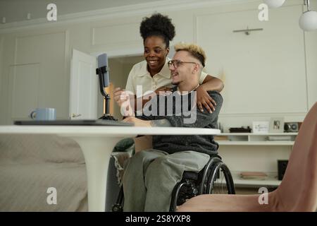 Freimütige Aufnahme einer fröhlichen Schwarzen Frau, die einen Mann sanft mit Rollstuhl umschließt und gemeinsam Inhalte für soziale Medien erstellt, während sie Spaß im Wohnzimmer hat Stockfoto
