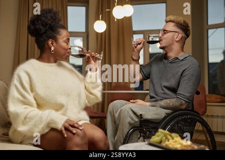 Seitenansicht eines Mannes, der im Rollstuhl köstliche Rotweine verkostet, bei einem romantischen Date mit einer charmanten Freundin im gemütlichen Wohnzimmer und einem gemeinsamen Moment zu Hause Stockfoto