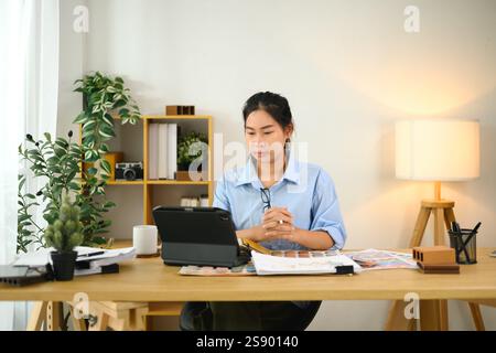 Lächelnder Innenarchitekt mit digitalem Tablet am Schreibtisch mit Farbmustern, Materialmustern und Designwerkzeugen Stockfoto