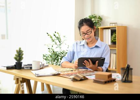 Lächelnder Innenarchitekt mit digitalem Tablet am Schreibtisch mit Farbmustern, Materialmustern und Designwerkzeugen Stockfoto