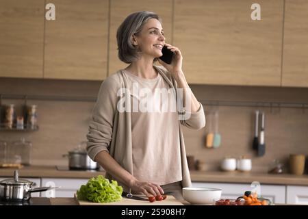 Reife Frau spricht auf dem Handy, abgelenkt von der Zubereitung von Salat Stockfoto