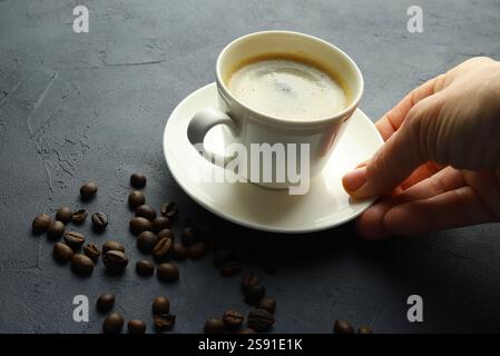 Weibliche Hand, die eine weiße Tasse Kaffee und Kaffeebohnen auf grauem Hintergrund hält. Direkt darüber. Stockfoto