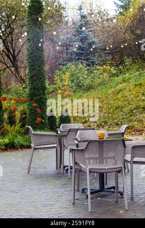 Ein leerer Caféplatz mit ein paar Tischen und Stühlen in einem grünen Park mit dekorativen Bäumen und einer Reihe von Lichtern, die sich über dem Park erstrecken. Stockfoto