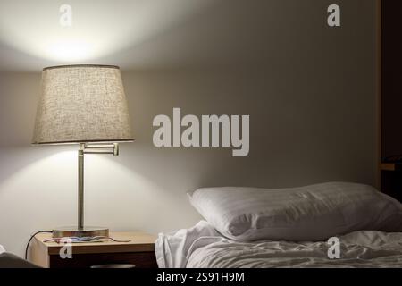 Ein Nachttisch mit Tischlampe beleuchtet ein leeres Bett mit zwei weißen Kissen. Stockfoto