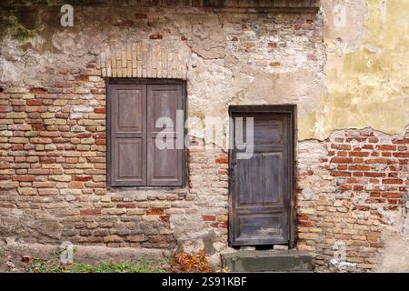 Ein Bruchstück eines alten Gebäudes mit verfallenem Putz, das eine Ziegelmauer mit einer Holztür und einem Fenster mit geschlossenen Flügeln freilegt. Stockfoto