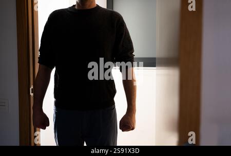 Ein dunkler Raum, eine Tür öffnet sich mit der Silhouette eines nicht erkennbaren Mannes, er steht in der Tür und sieht sein Opfer an. Fokus auf Schatten. Stockfoto
