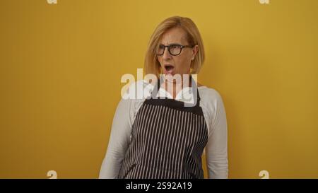 Frau mittleren Alters mit kurzen blonden Haaren in gestreifter Schürze, überraschend auf gelbem Hintergrund Stockfoto