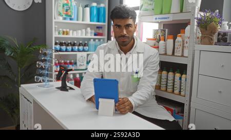 Schöner hispanischer Apotheker Mann, der Tablette in einer gut sortierten Apotheke mit Regalen für Medizin und Gesundheitsprodukte verwendet Stockfoto