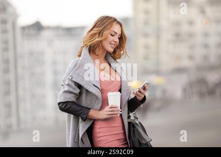 Eine Geschäftsfrau, die E-Mails auf ihrem Handy liest und eine Kaffeetasse in der Hand hält, vor einem städtischen Hintergrund Stockfoto