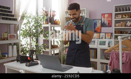 Junger Mann in einem Wohnkultur-Laden, der mit einem Laptop hinter einer Theke klatscht, umgeben von Pflanzen und verschiedenen Dekorationen. Stockfoto