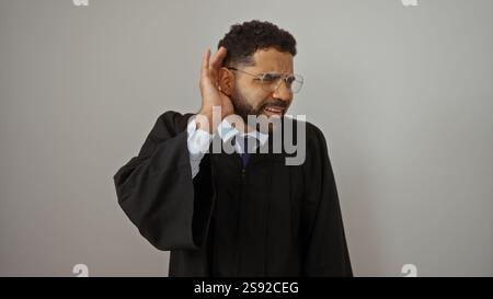 Ein schöner junger Mann im Abschlusskleid posiert isoliert vor weißem Hintergrund und hält eine aufmerksame Hand-Ohr-Geste aufrecht Stockfoto