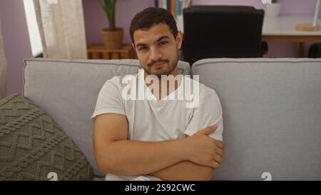 Junger hispanischer Mann, der entspannt auf einer Couch im Wohnzimmer eines Hauses sitzt, mit überkreuzten Armen und einem neutralen Ausdruck Stockfoto