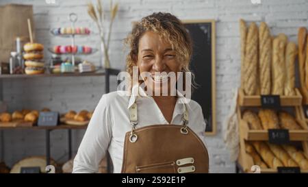 Frau lächelt in einer Bäckerei, umgeben von Brot und Gebäck, trägt eine Schürze, mittleres Alter, Inneneinrichtung, warme Atmosphäre, reife, weibliche Ladeninhaberin, Stockfoto