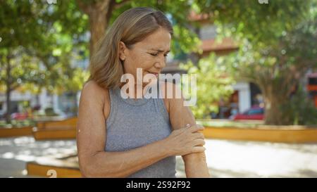 Frau kratzt sich den Arm in einer Parklandschaft im Freien, mit alten Bäumen und einer lebhaften Stadtumgebung im Hintergrund, die Unannehmlichkeiten oder alles repräsentiert Stockfoto